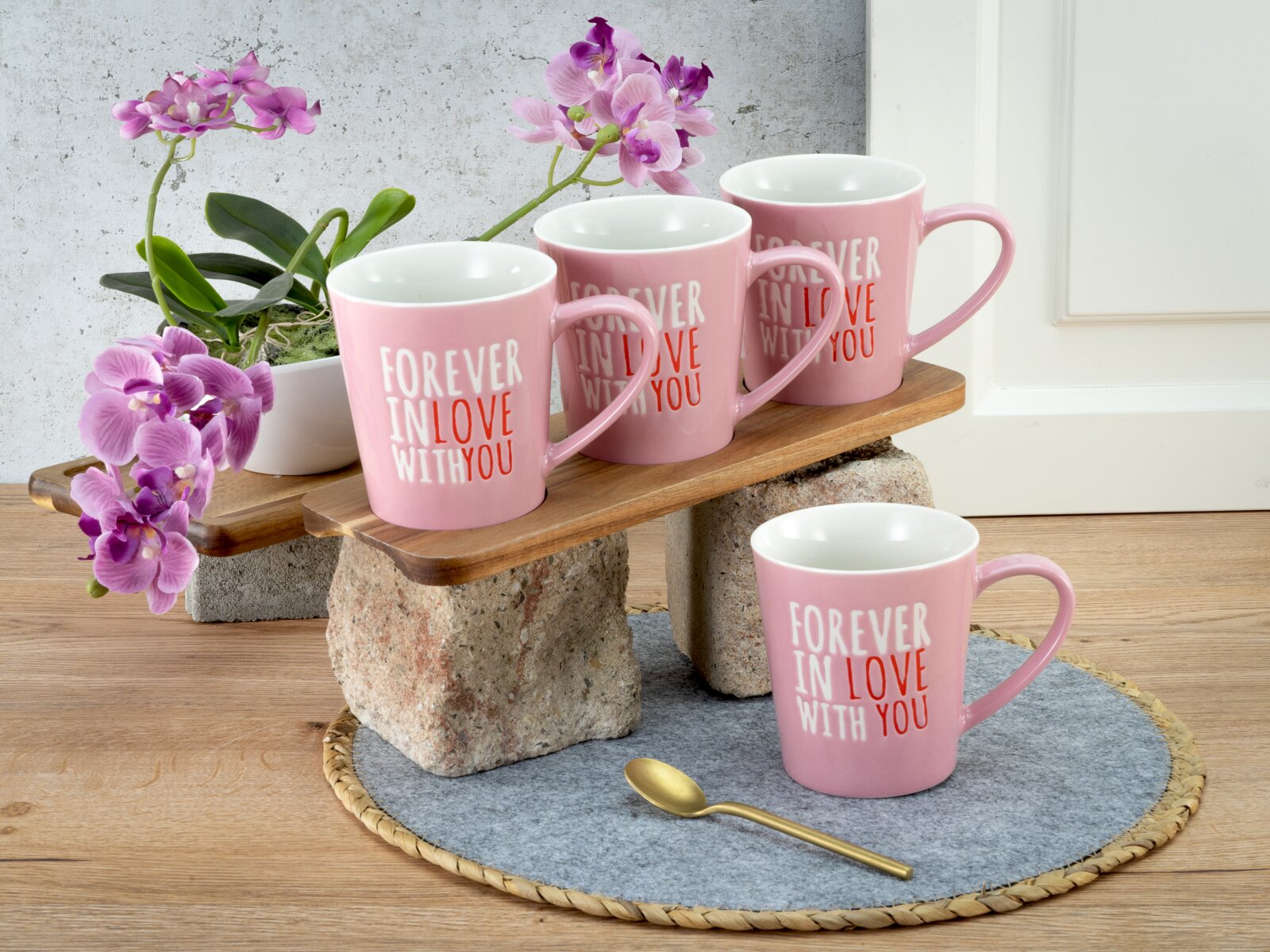 Vier rosafarbene Keramiktassen mit der Aufschrift „FOREVER IN LOVE WITH YOU“ auf Holzbrett-Ständer, daneben Orchideen und goldener Löffel Perspektive: leicht erhöhte Frontansicht.