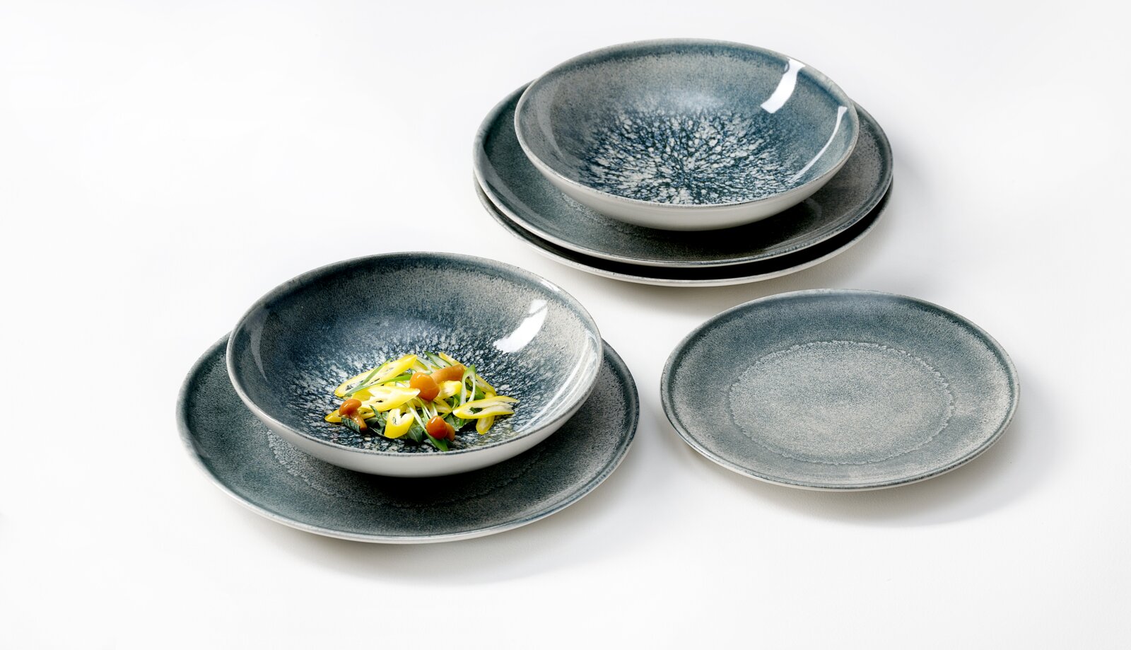 Blauwgrijs keramisch servies met organisch gespikkeld glazuur, bestaande uit kommen en borden, schuin van boven gefotografeerd op een witte achtergrond.
