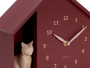 Nahaufnahme einer burgunderroten Wanduhr in Hausform mit einer kleinen Katzenfigur im Inneren. Die Uhr hat beige Zeiger und Ziffern. Perspektive von vorne leicht seitlich.