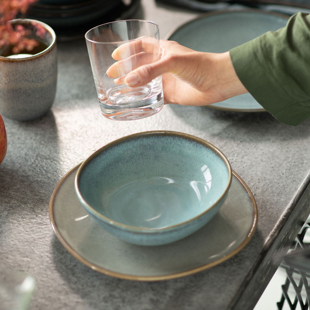 Türkise Müslischale Lave 380ml auf einem passenden Teller, von oben betrachtet, neben einem Glas in einer Hand.