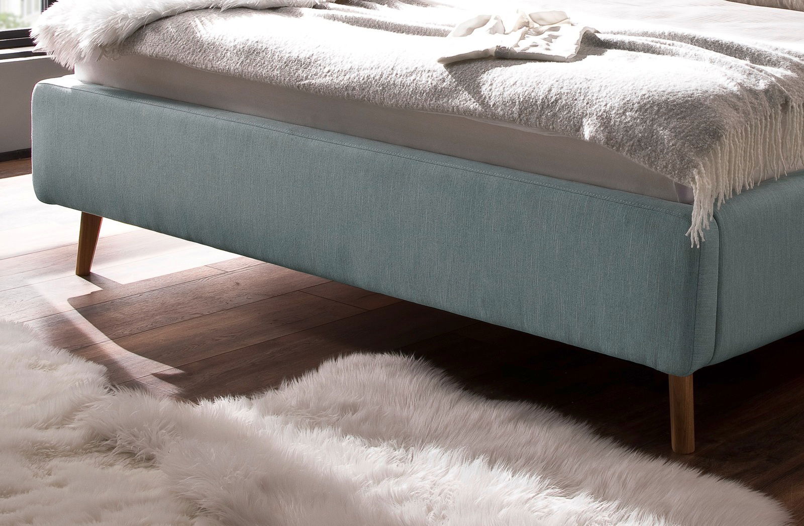 Seitliche Perspektive eines Polsterbetts in hellem Türkis mit Holzfüßen auf einem Holzboden und einem weißen Teppich.