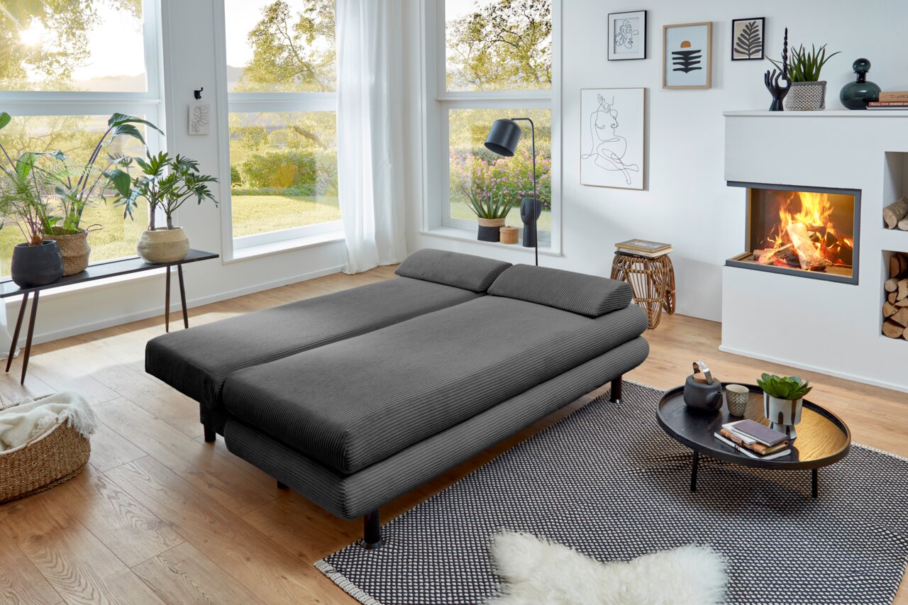 Graues Funktionssofa in einem modernen Wohnzimmer mit Blick auf den Garten, umgeben von Pflanzen und einem Kamin.