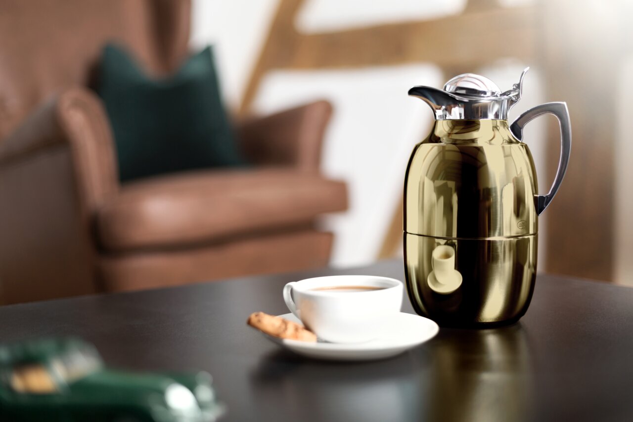 Goldene Isolierkanne Juwel auf einem Tisch, neben einer Tasse Kaffee und einem Keks, im Hintergrund ein unscharfer Sessel. Perspektive: seitlich.