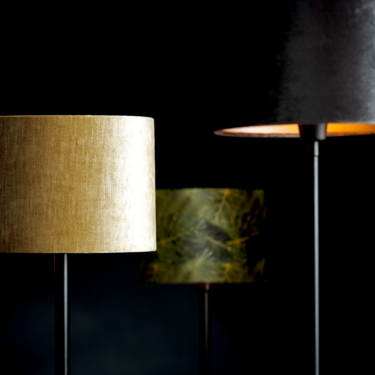 Drei Lampenschirme in verschiedenen Farben und Mustern, im Vordergrund ein goldener Zylinder-Lampenschirm, seitliche Perspektive.
