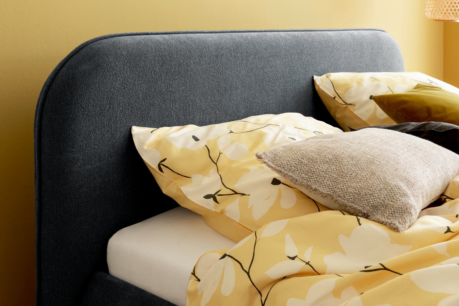 Nahaufnahme eines Polsterbettgestells mit grauem Kopfteil, gelber Bettwäsche mit Blumenmuster und mehreren Kissen, seitliche Perspektive.