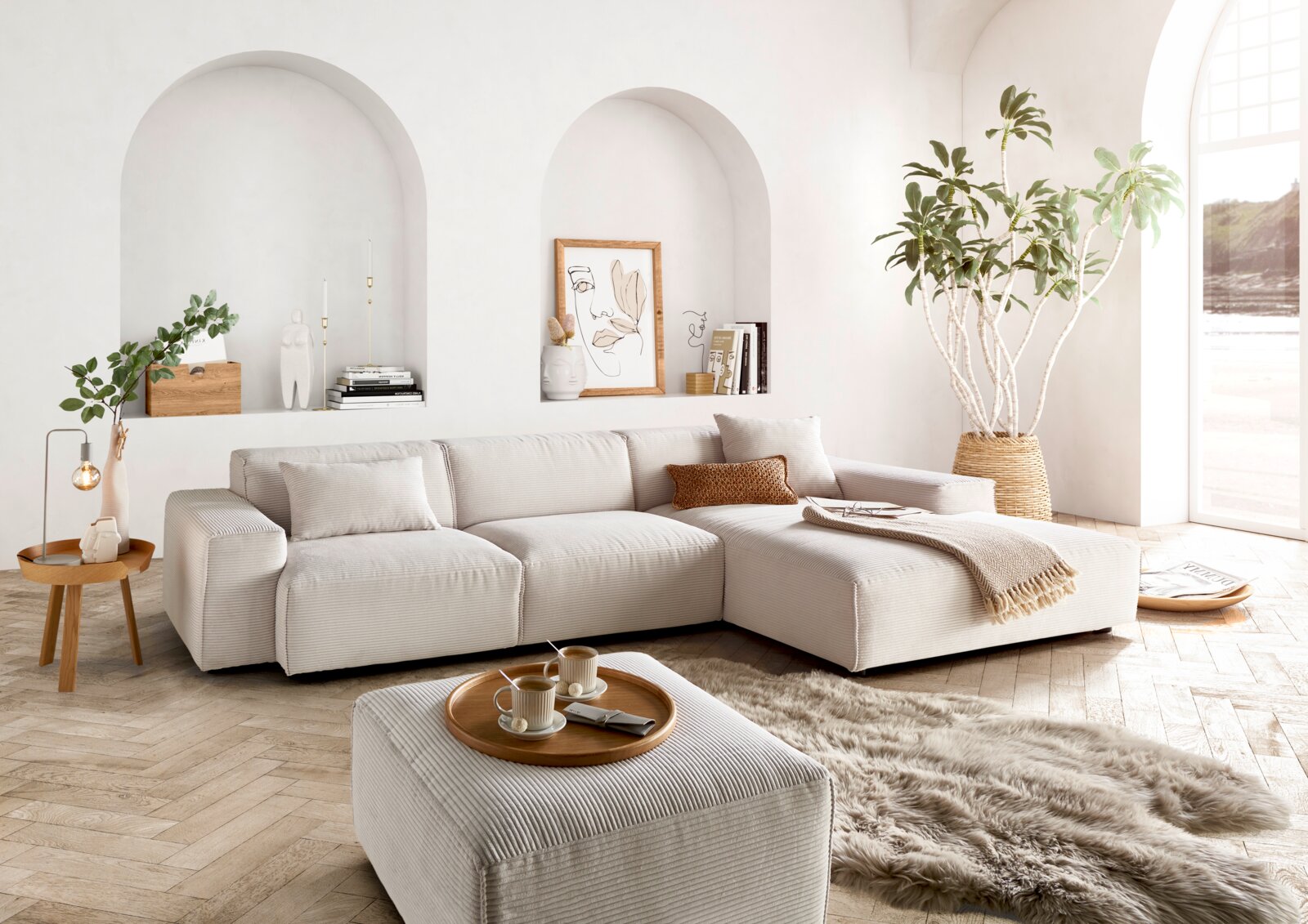 Lounge Handmade Ecksofa LH-EDINA Moderne, helle Wohnzimmerecke mit einer großen, beigen Rundecke und einem passenden Hocker, umgeben von stilvollen Dekorationen und Pflanzen. Perspektive von vorne.