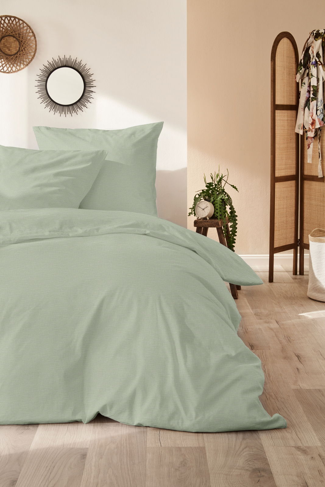 Bettwäsche-Set in sanftem Grün, bestehend aus einem Bettbezug und zwei Kissenbezügen, auf einem Bett in einem stilvoll eingerichteten Schlafzimmer, aus der Vorderansicht fotografiert.