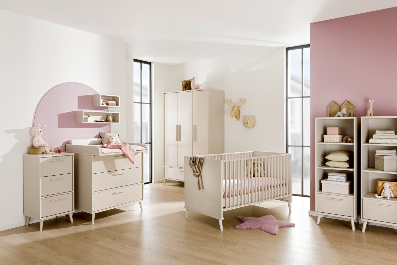 Helles Babyzimmer mit weißem Babybett, Kleiderschrank, Wickelkommode und Regalen in skandinavischem Stil, seitliche Perspektive in einem sonnigen Raum mit rosa Akzentwand.