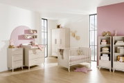 Helles Babyzimmer mit weißem Babybett, Kleiderschrank, Wickelkommode und Regalen in skandinavischem Stil, seitliche Perspektive in einem sonnigen Raum mit rosa Akzentwand.