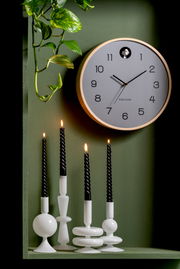 Moderne Wanduhr mit hellem Holzrahmen und klaren Ziffern auf grünem Hintergrund, umgeben von Pflanzen und brennenden schwarzen Kerzen. Frontalansicht.