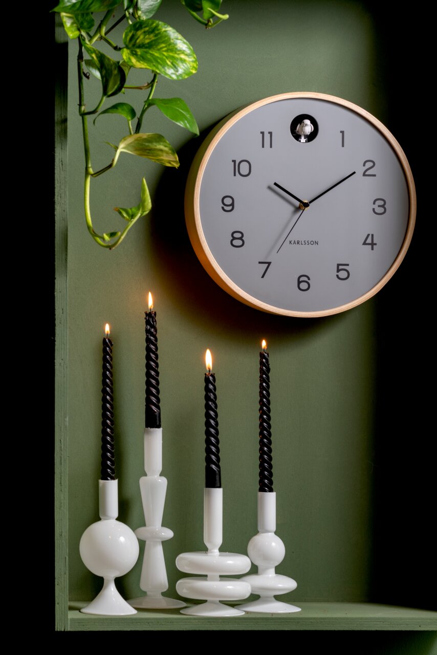 Moderne wandklok met een lichte houten lijst en duidelijke cijfers op een groene achtergrond, omringd door planten en brandende zwarte kaarsen. Vooraanzicht.