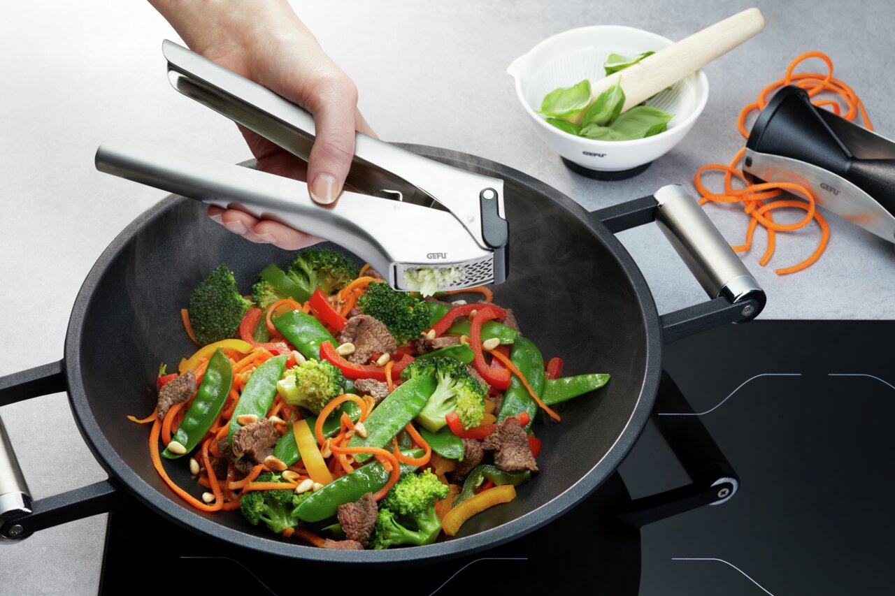 Knoblauchpresse GARLICO in der Hand, die frischen Knoblauch über einer Pfanne mit Gemüse und Fleisch presst, seitliche Perspektive.