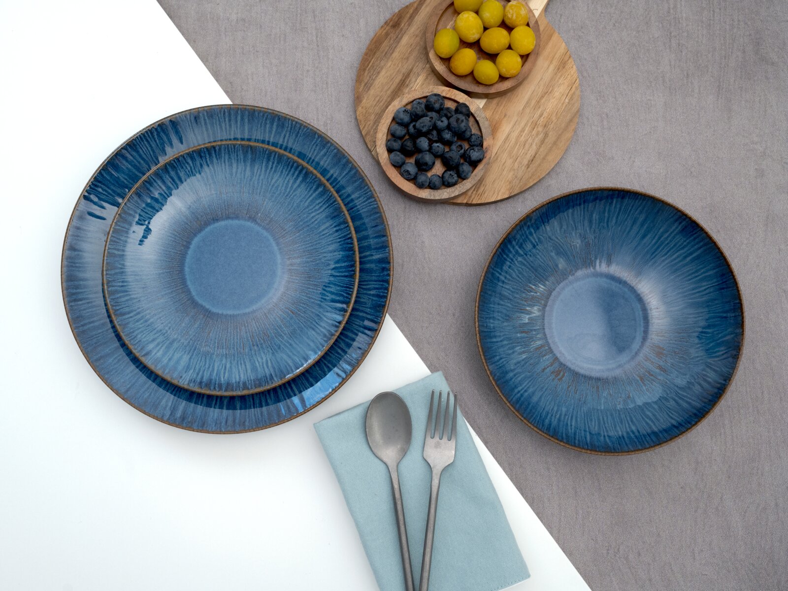 Drei blaue Keramik-Teller mit reaktivem Glasurverlauf und braunem Rand, arrangiert auf grauem und weißem Hintergrund mit Holzschalen voller Beeren sowie Besteck auf Serviette, Draufsicht (Flatlay)