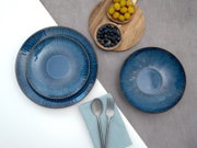 Drei blaue Keramik-Teller mit reaktivem Glasurverlauf und braunem Rand, arrangiert auf grauem und weißem Hintergrund mit Holzschalen voller Beeren sowie Besteck auf Serviette, Draufsicht (Flatlay)