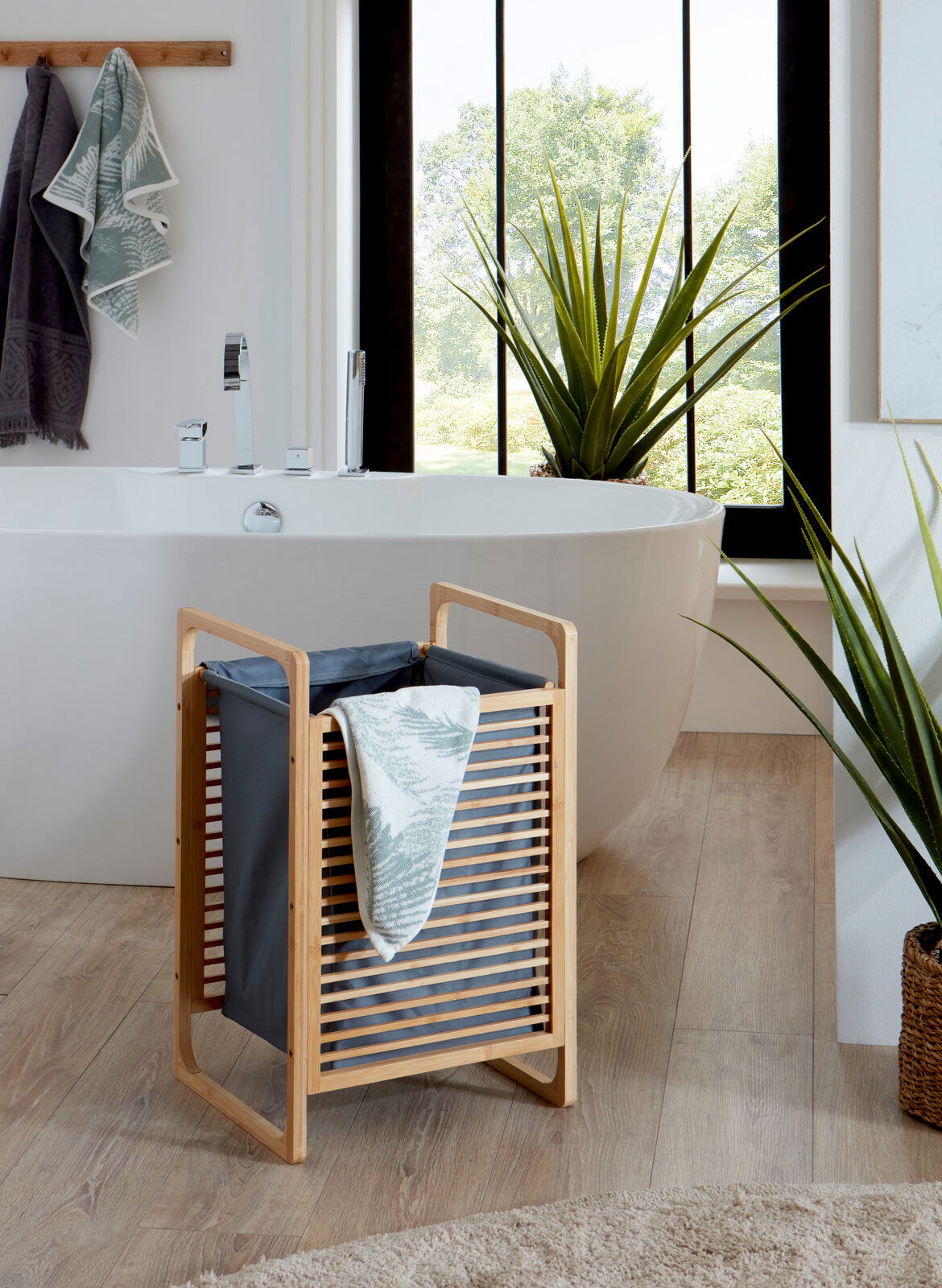 Bambus-Wäschekorb mit grauem Stoffeinsatz und Handtuch, im Badezimmer vor freistehender Badewanne; Perspektive: frontale, leicht erhöhte Ansicht.