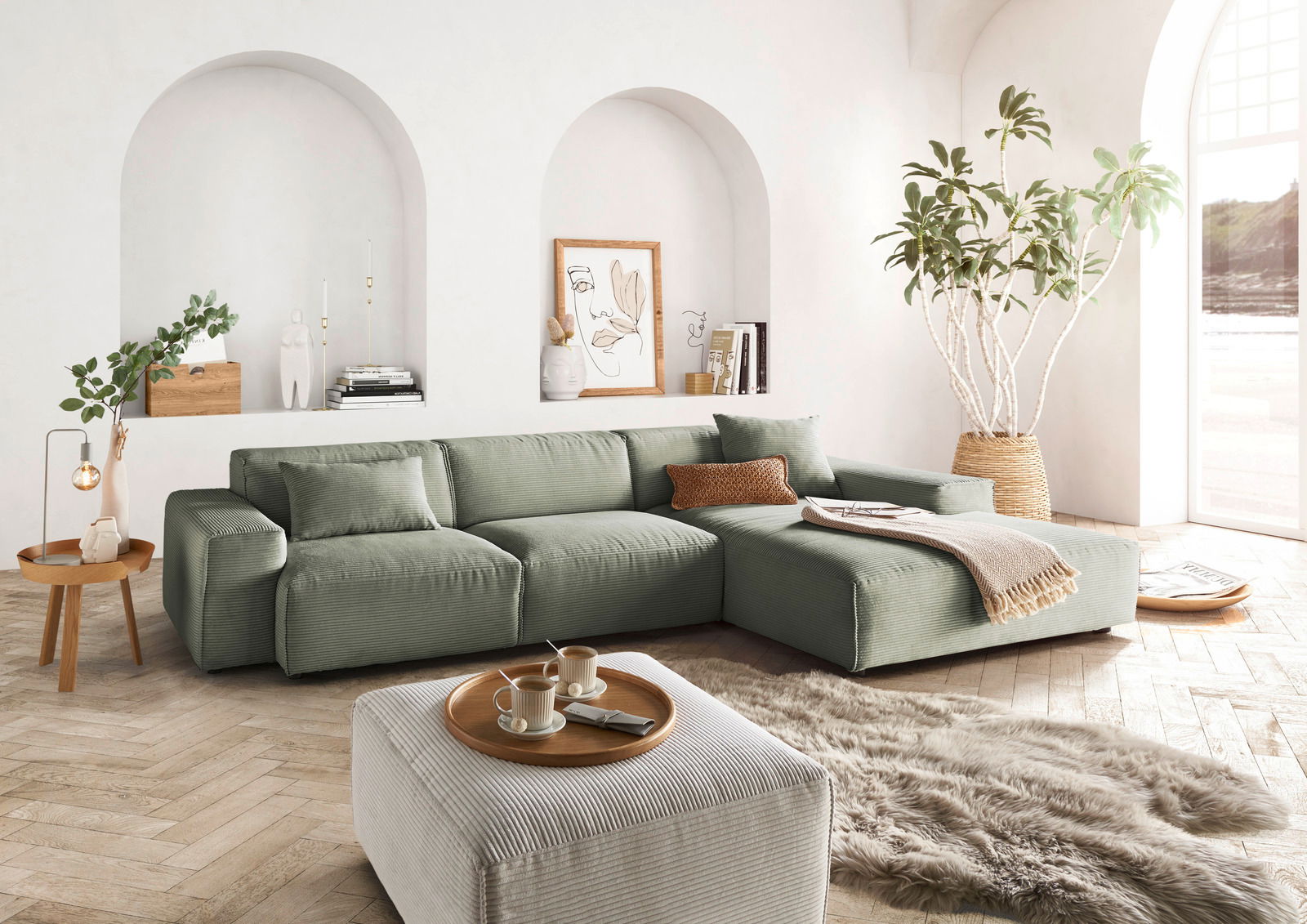 Lounge Handmade Hoekbank LH-EDINA Grüne Ecksofa in einem hellen Wohnzimmer mit großen Fenstern, Pflanzen und dekorativen Elementen, aus der Perspektive von vorne links.