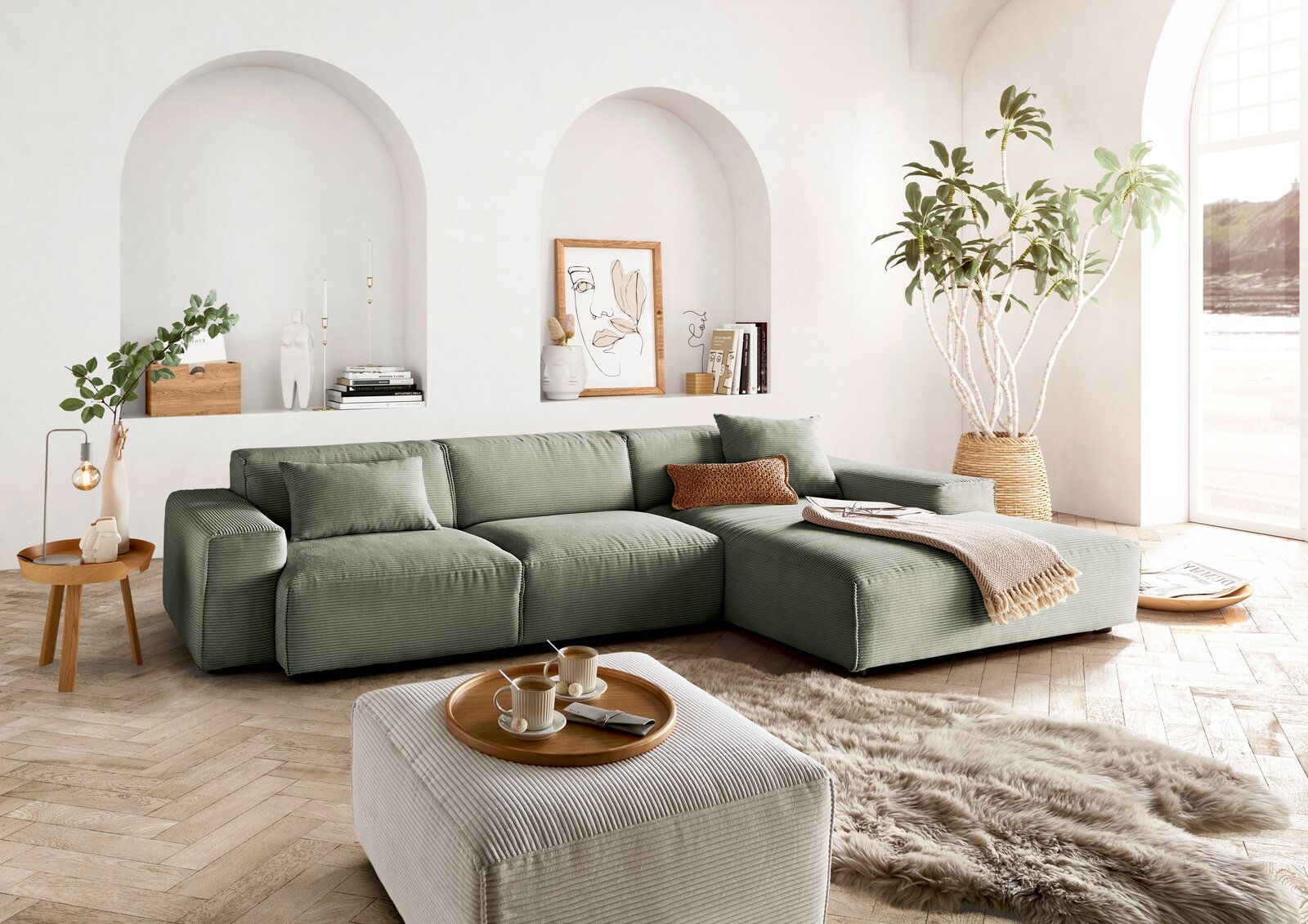 Lounge Handmade Ecksofa LH-EDINA Grüne Ecksofa in einem hellen Wohnzimmer mit großen Fenstern, Pflanzen und dekorativen Elementen, aus der Perspektive von vorne links.