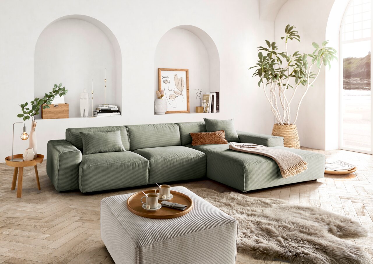 Lounge Handmade Ecksofa LH-EDINA Grüne Ecksofa in einem hellen Wohnzimmer mit großen Fenstern, Pflanzen und dekorativen Elementen, aus der Perspektive von vorne links.