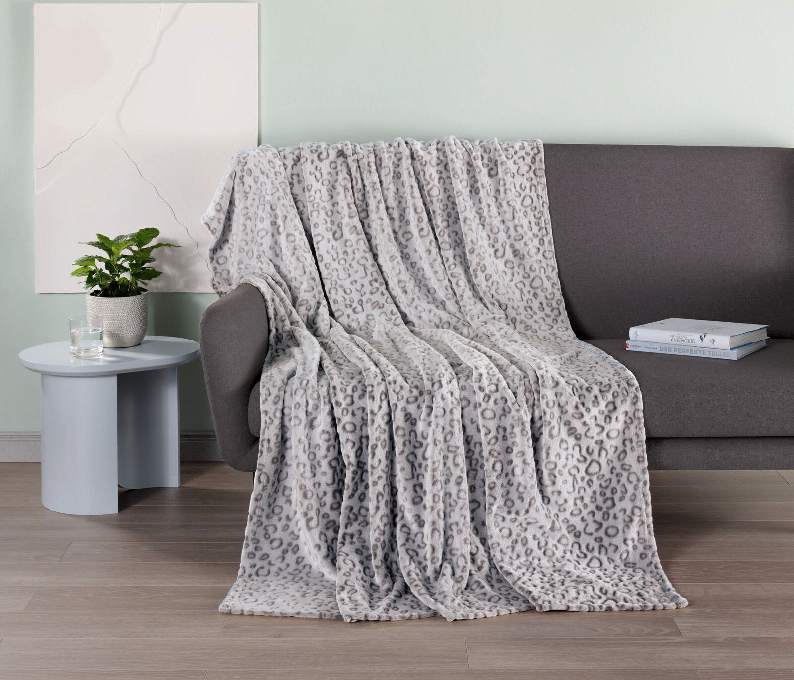Graue Wohndecke mit Leopardenmuster, drapiert über einem dunklen Sofa, mit einem Beistelltisch und Pflanzen im Hintergrund, aus seitlicher Perspektive aufgenommen.