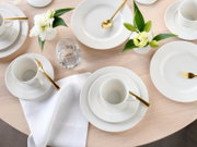 Elegantes weißes Porzellan-Geschirrset mit Tassen, Tellern und Schalen auf hellem Holztisch, ergänzt durch goldfarbenes Besteck, Wasserglas, weiße Serviette und Blumenarrangements – Draufsicht von oben