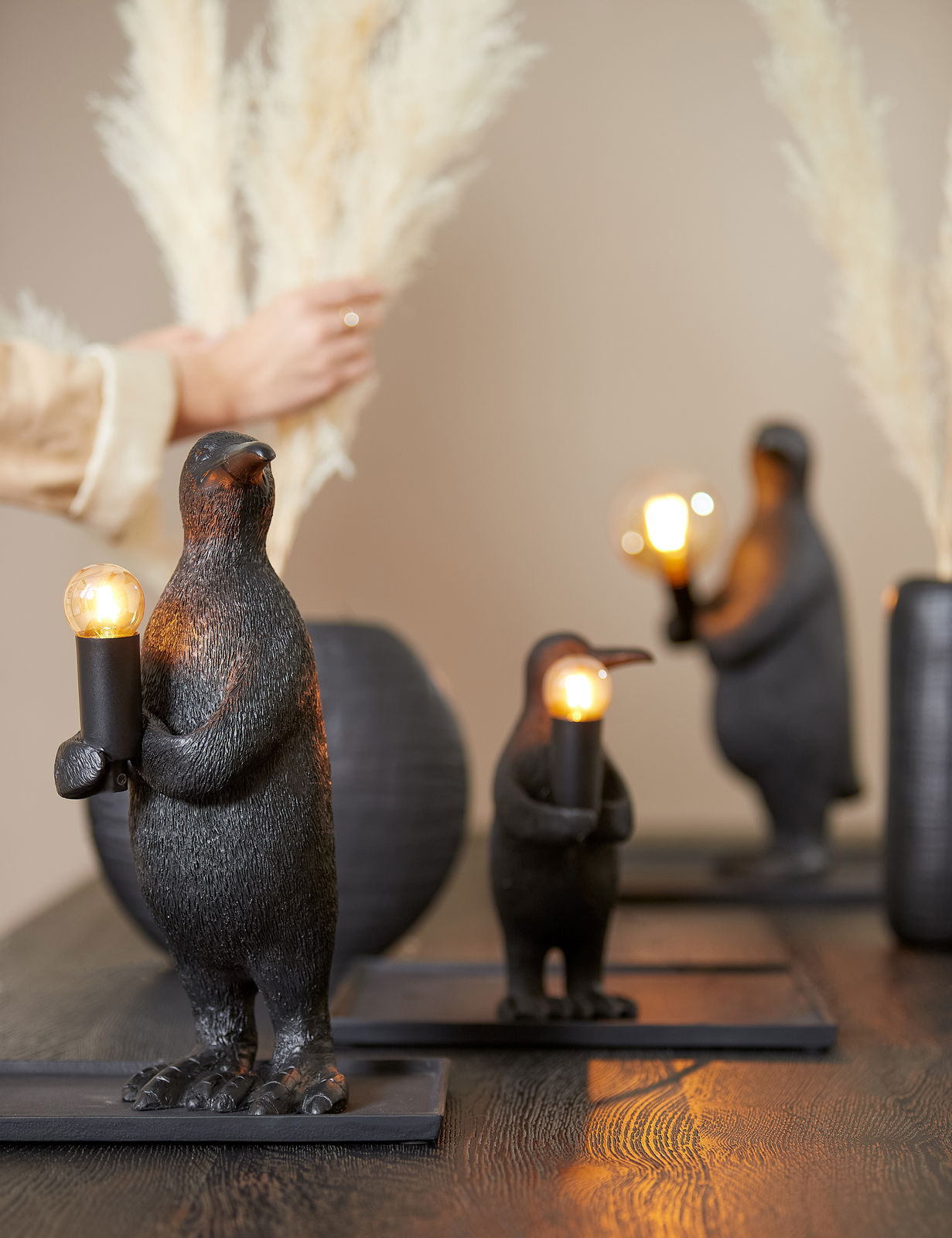 Light&Living Tischleuchte PENGUIN Drei Pinguin-förmige Tischleuchten aus schwarzem Material, die auf einem Tisch stehen. Jede Leuchte hält eine Glühbirne in den Flügeln. Die Perspektive zeigt die Leuchten von vorne, mit einer unscharfen Hand im Hintergrund, die Pampasgras arrangiert.
