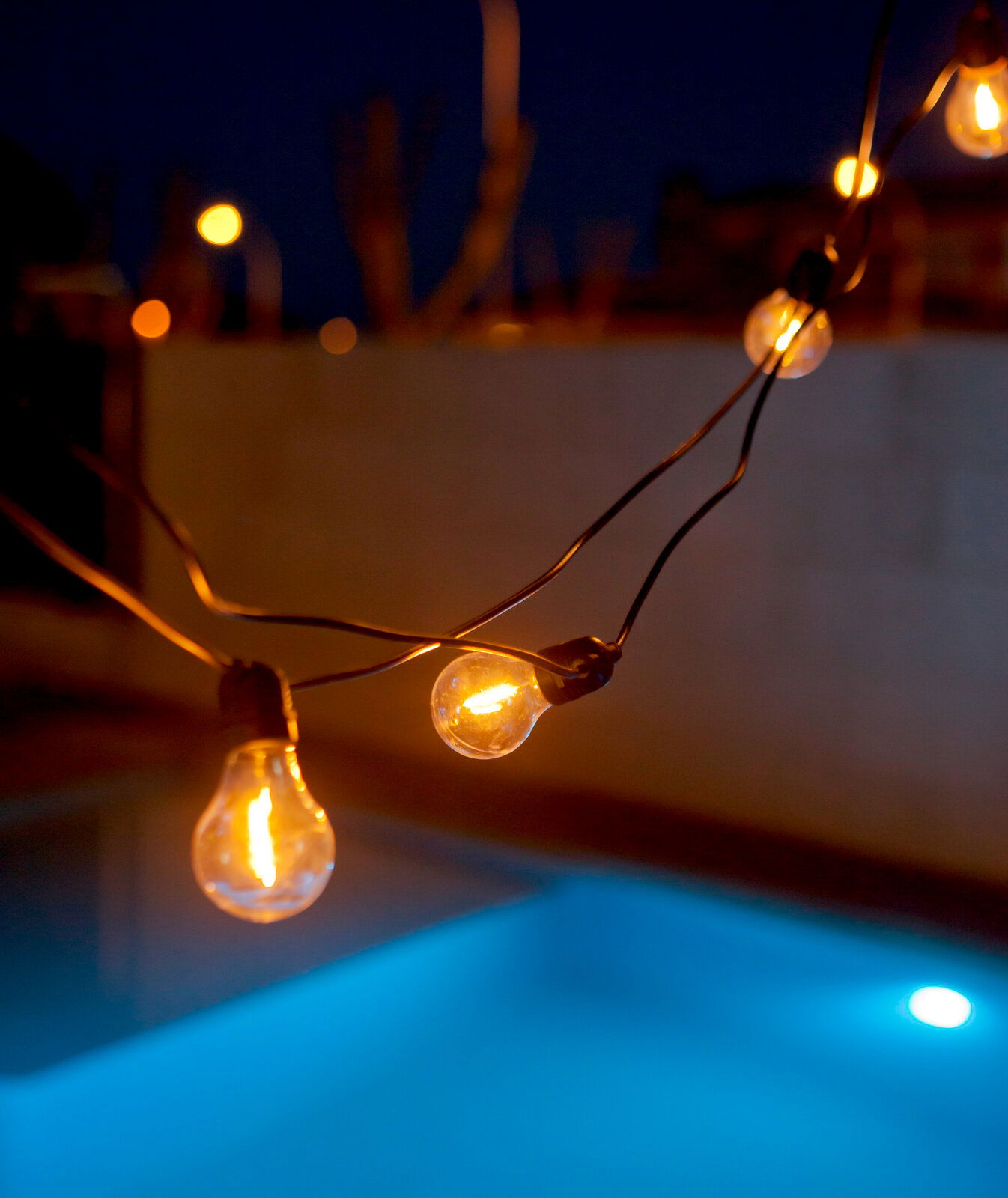 Nahaufnahme aus leicht seitlicher Perspektive: warm leuchtende Lichterkette mit Glühbirnen über einem Pool bei Dämmerung, Hintergrund unscharf