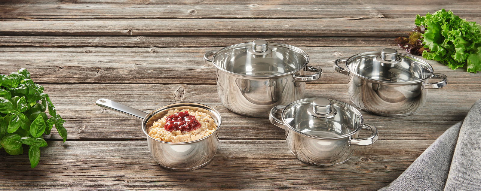 Kochset aus Edelstahl, bestehend aus vier Töpfen mit Deckeln, auf einer Holzoberfläche von oben fotografiert. Frische Kräuter und Salatblätter sind im Hintergrund zu sehen.