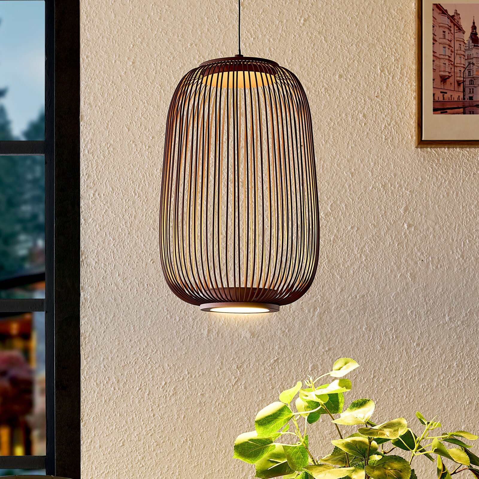 Hängelampe mit ovalem, käfigartigem Lampenschirm aus dunklem Holz/Lamellen und warmem Licht, frontal in leicht seitlicher Innenraumansicht vor heller Wand