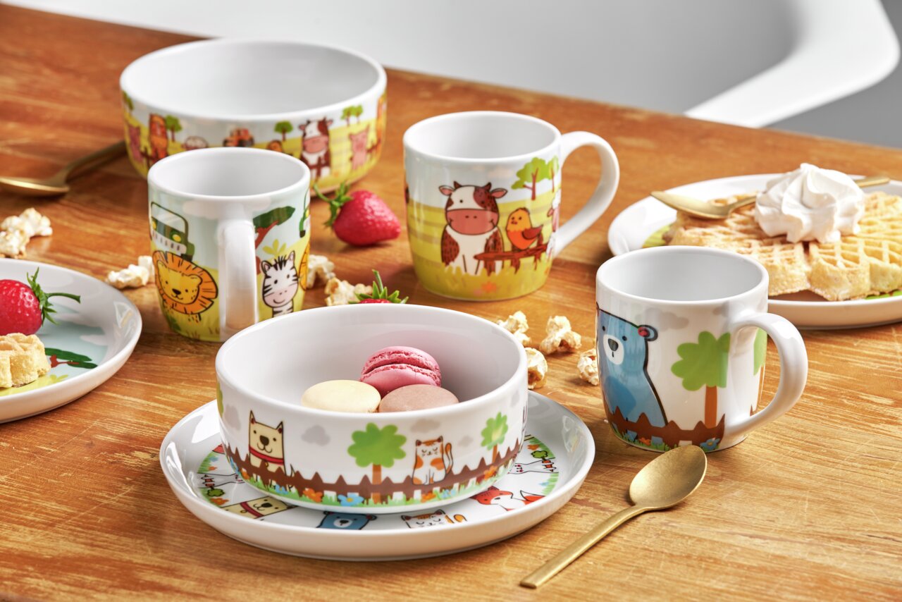 Kinderset mit bunten Tiermotiven, bestehend aus Tassen, Schalen und Tellern, auf einem Holztisch arrangiert. Perspektive von schräg oben.