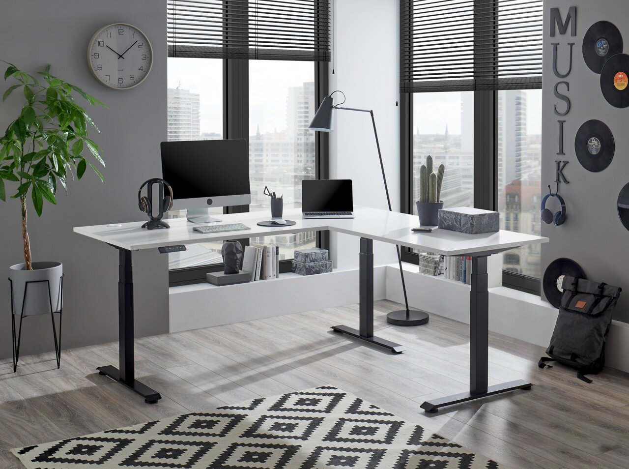 Moderner E-Eckschreibtisch in einem stilvollen Büro mit Blick auf die Stadt, ausgestattet mit Computer, Laptop und Pflanzen, aus der Perspektive von vorne links.