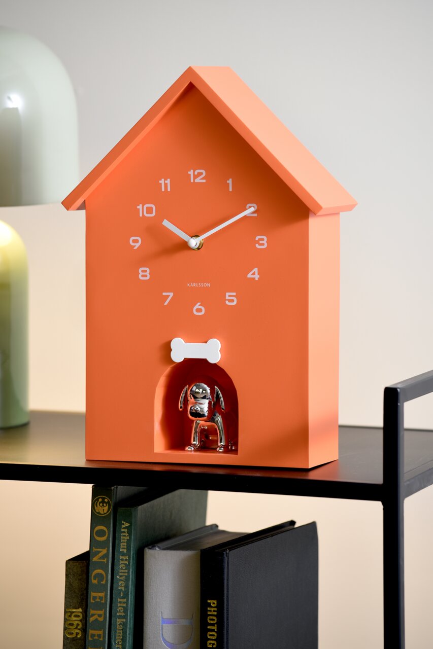 Orangefarbene Wanduhr in Form eines Hauses mit weißen Ziffern und Zeigern, darunter eine silberne Hundefigur in einer kleinen Nische. Perspektive von vorne leicht seitlich.