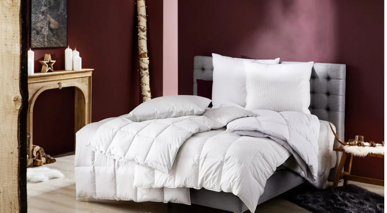 Gemütliches Schlafzimmer mit einem grauen, gepolsterten Bett, das mit einer weißen Daunendecke und Kissen ausgestattet ist. Die Perspektive zeigt das Bett von der Seite, umgeben von einer warmen, einladenden Dekoration mit Kerzen und Holzakzenten.