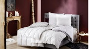 Gemütliches Schlafzimmer mit einem grauen, gepolsterten Bett, das mit einer weißen Daunendecke und Kissen ausgestattet ist. Die Perspektive zeigt das Bett von der Seite, umgeben von einer warmen, einladenden Dekoration mit Kerzen und Holzakzenten.