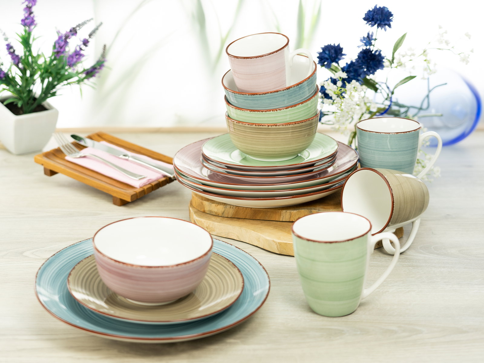Kombiservice 16-tlg in verschiedenen Pastellfarben, bestehend aus Tellern, Schalen und Tassen, auf einem Tisch arrangiert. Perspektive von vorne.