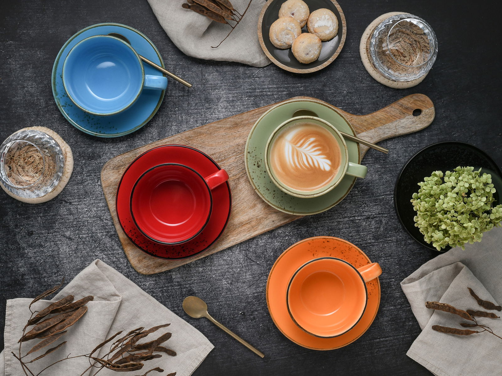 Vogelperspektive auf ein Set von bunten Cappuccinotassen in Blau, Rot, Grün und Orange, arrangiert auf einem dunklen Tisch mit einem Holzbrett, Gebäck und Dekoration.