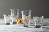 Vier Gläser aus dem 12-teiligen Becherset TWIST, auf einem Tisch arrangiert, mit einem Glas gefüllt mit Wasser und Orangenscheiben, seitliche Perspektive.