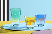Drei bunte Trinkgläser in den Farben Grün, Gelb und Blau auf einem farbenfrohen Hintergrund, mit einem Glas gefüllt mit einem Getränk und Orangenscheiben, aus der Seitenperspektive fotografiert.