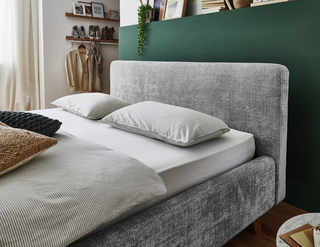 Graues Polsterbettgestell mit Kopfteil, seitliche Perspektive, dekoriert mit Kissen und Decken in einem gemütlichen Schlafzimmer.