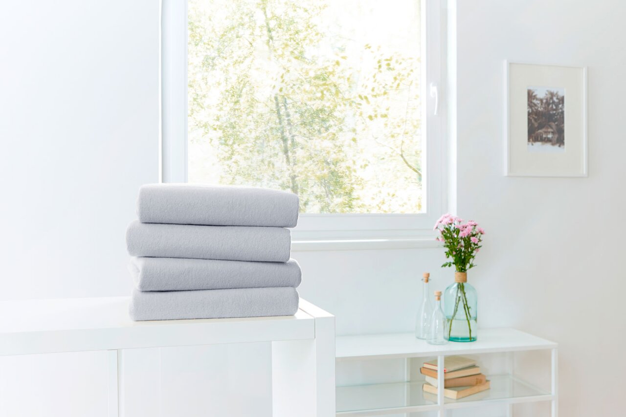 Novel Spannbetttuch TEDDY-FLAUSCH Ein Stapel grauer Spannbetttücher auf einem weißen Tisch vor einem Fenster mit Blick ins Grüne, seitliche Perspektive.