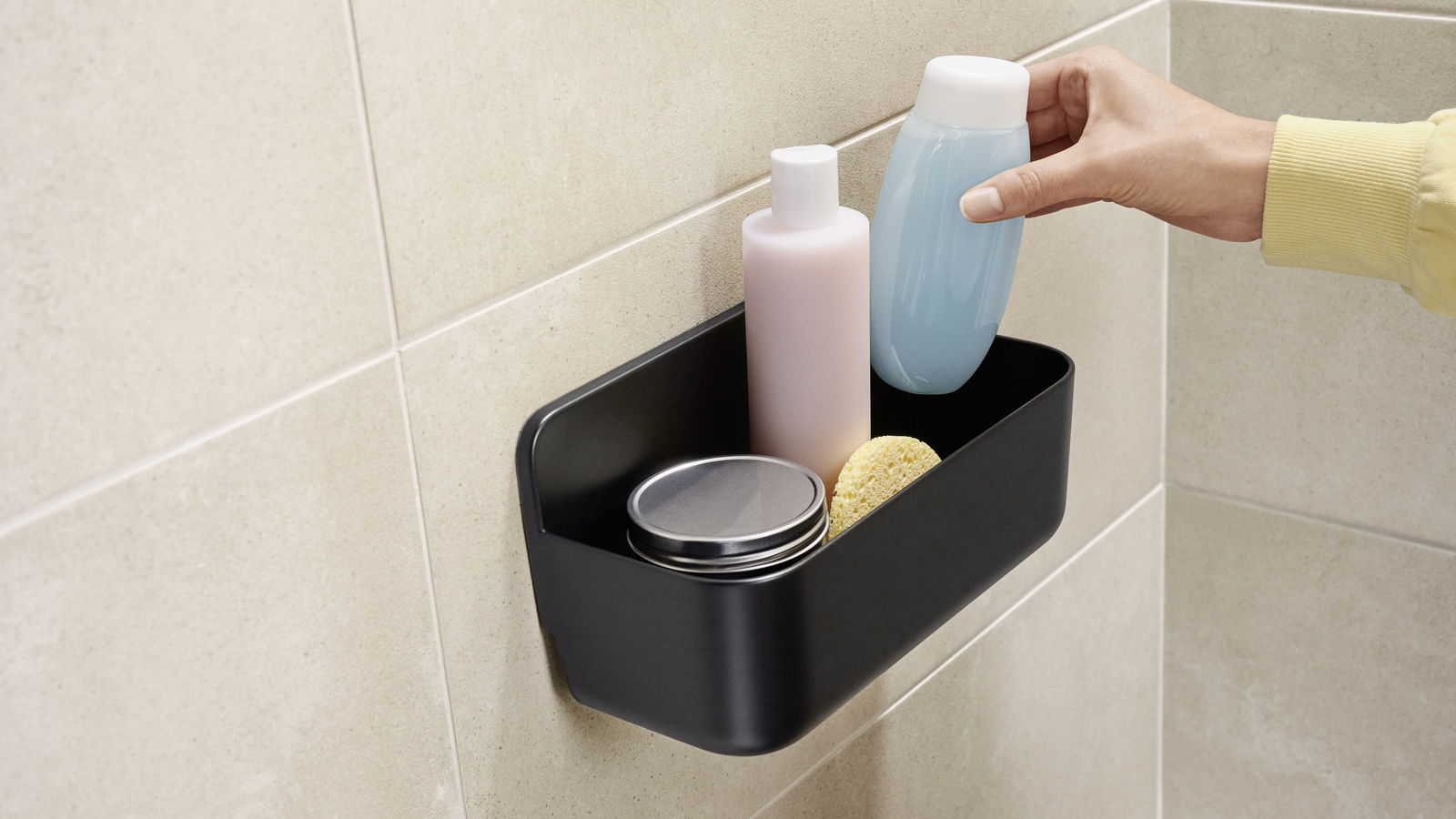 Schwarzer Duschkorb an der Wand montiert, gefüllt mit Pflegeprodukten und einer Hand, die eine Flasche entnimmt. Seitliche Perspektive.