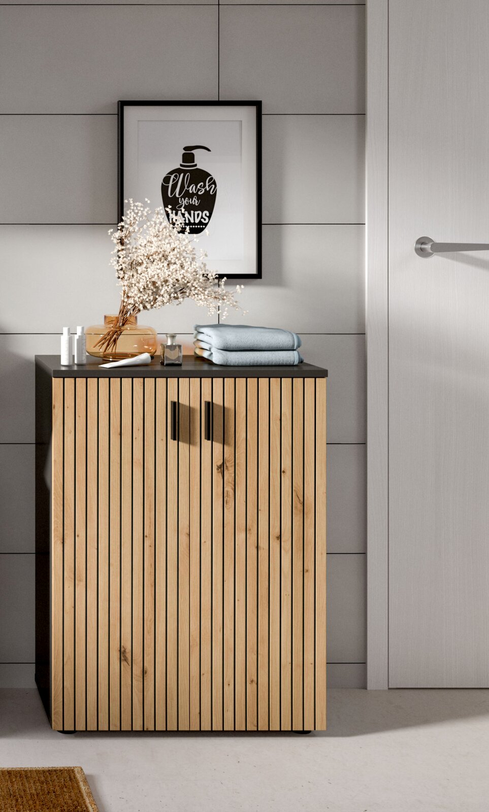 Modernes Badmöbel/Unterschrank aus hellem Holz mit vertikaler Lamellenfront und schwarzer Ablageplatte, darauf Handtücher und Deko, vor grauer Fliesenwand mit gerahmtem „Wash your hands“-Poster; Frontansicht auf Augenhöhe.