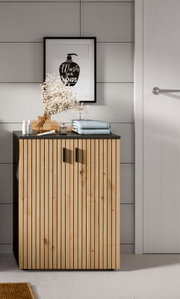 Modernes Badmöbel/Unterschrank aus hellem Holz mit vertikaler Lamellenfront und schwarzer Ablageplatte, darauf Handtücher und Deko, vor grauer Fliesenwand mit gerahmtem „Wash your hands“-Poster; Frontansicht auf Augenhöhe.