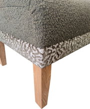 Close-up van de voorste hoek van een gestoffeerde stoel met grijze bekleding, een zijstuk met patroon en een houten poot, schuin van bovenaf gefotografeerd