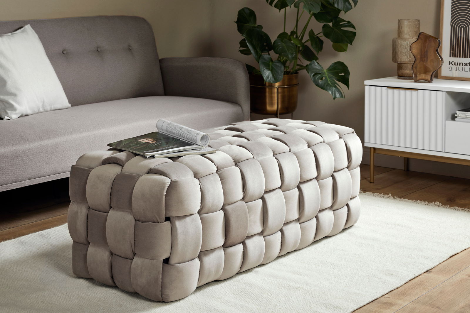 Beiger, gepolsterter Hocker in geflochtener Optik, seitliche Perspektive, platziert in einem modernen Wohnzimmer mit Sofa und Beistelltisch.