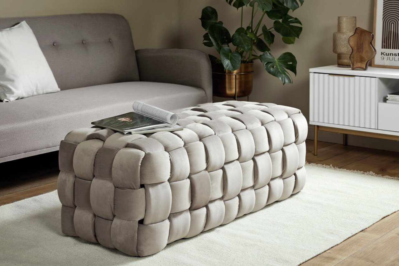 Beiger, gepolsterter Hocker in geflochtener Optik, seitliche Perspektive, platziert in einem modernen Wohnzimmer mit Sofa und Beistelltisch.