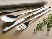 Elegantes Besteck-Set mit gehämmerter Oberfläche, bestehend aus Löffel, Messer und Gabel, auf einem Holztisch mit Kräutern und Stoffserviette, aus der Vogelperspektive fotografiert.