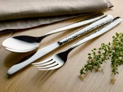 Elegante bestekset met gehamerd oppervlak, bestaande uit een lepel, mes en vork, op een houten tafel met kruiden en een stoffen servet, gefotografeerd vanuit vogelperspectief.