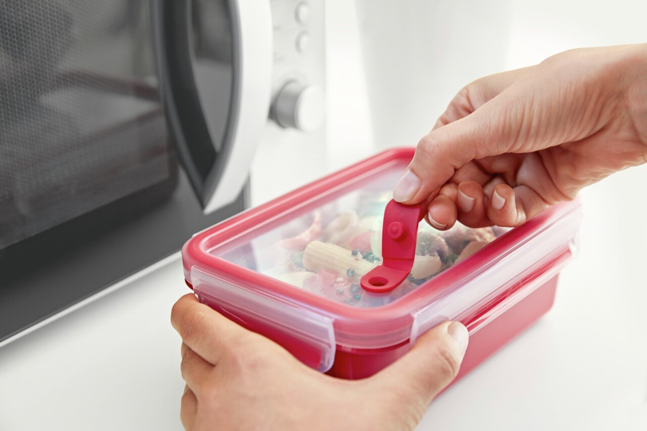 Rote Mikrowellendose mit transparentem Deckel, die von einer Hand geöffnet wird, vor einer Mikrowelle.