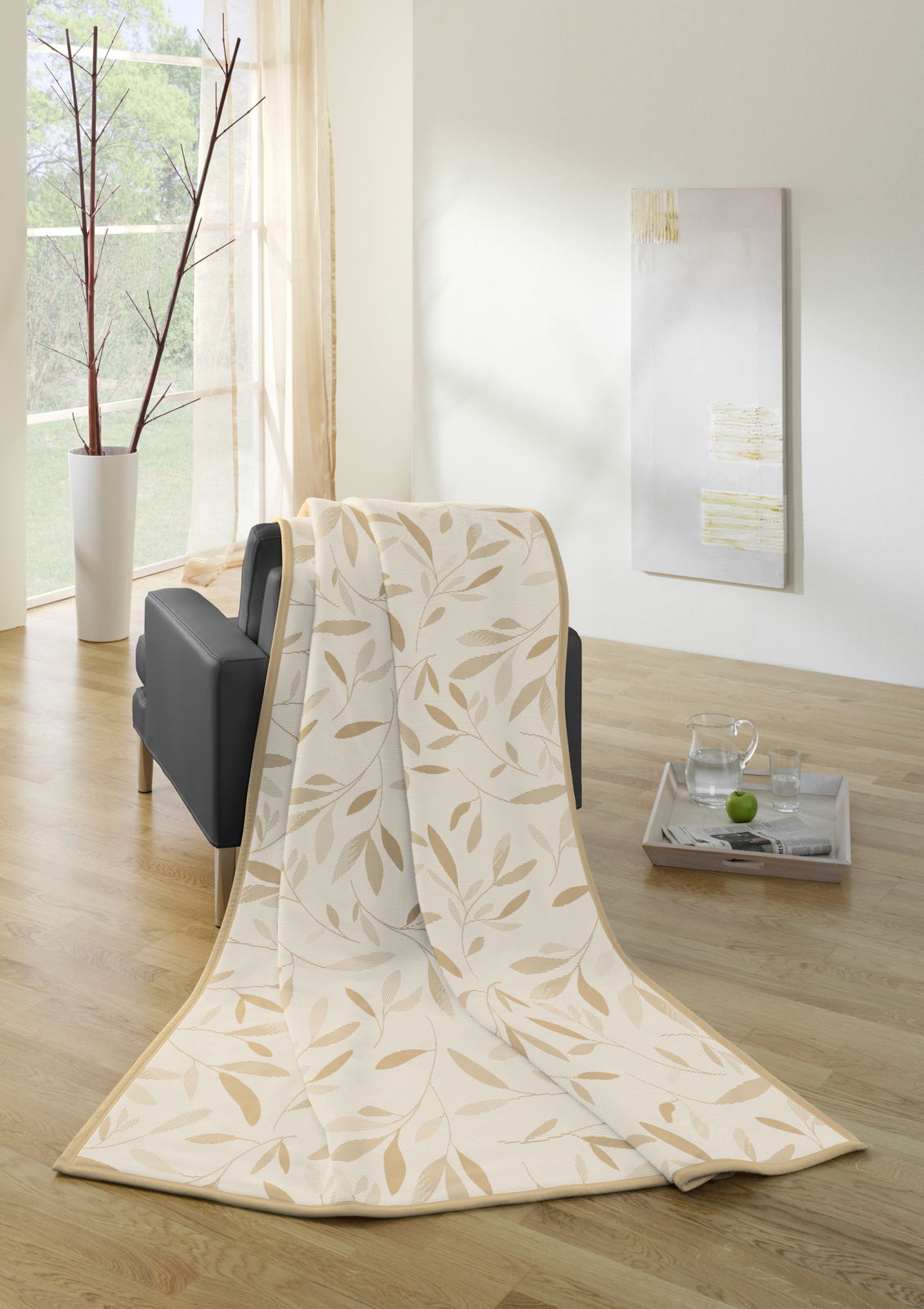 biederlack Wohndecke COTTON HOME Wohndecke mit Blättermuster in Beige, drapiert über einem schwarzen Sofa in einem hellen Raum mit Holzfußboden, seitliche Perspektive.