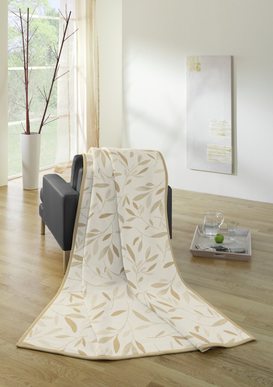 Wohndecke mit Blättermuster in Beige, drapiert über einem schwarzen Sofa in einem hellen Raum mit Holzfußboden, seitliche Perspektive.